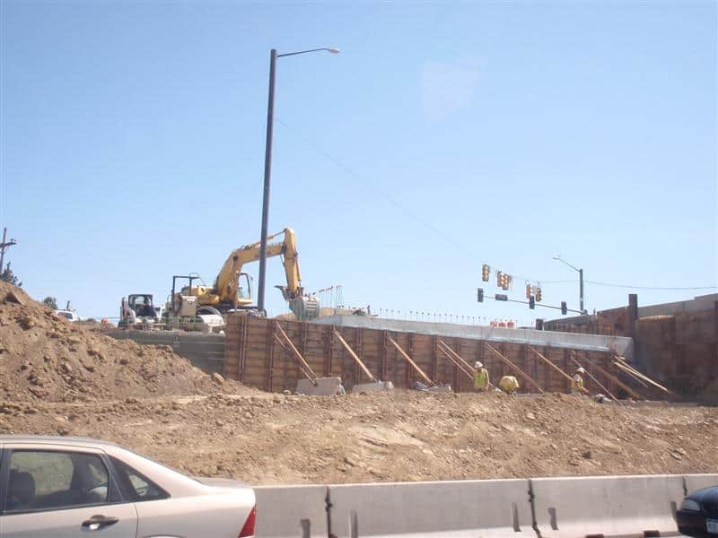 Yellow excavator digs and positions materials beside a wooden formwork wall while three construction workers brace it; dirt piles, concrete barriers, roadside cars and traffic lights frame a sunny intersection.