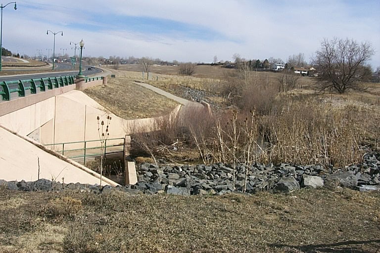 Concrete roadway bridge crosses a rocky drainage channel, directing runoff beneath; adjacent grassy fields and leafless shrubs stretch toward distant houses and street lamps under a pale winter sky.
