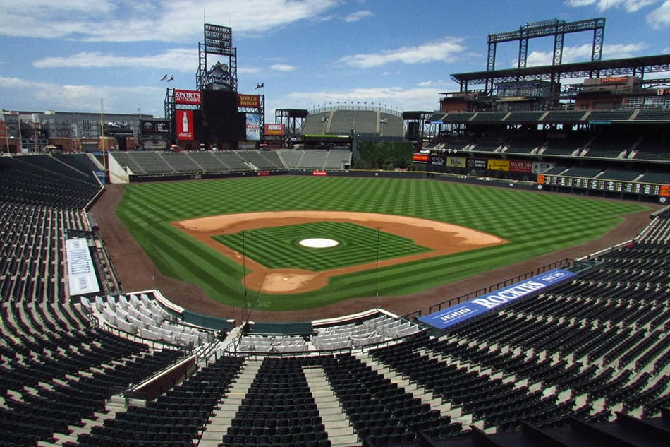 Baseball field — empty diamond and striped outfield grass resting between seating tiers in a large open-air stadium under a blue sky.

Text visible: "ROCKIES", "SPORTS AUTHORITY", "WELLS FARGO", "Coca-Cola".