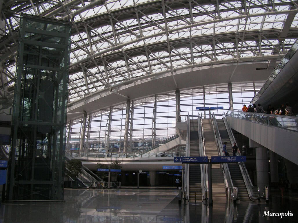 Central pair of escalators (object) ferry a few people upward (action) inside a vast glass-and-steel airport atrium with arched skylight, glass elevator shaft and reflective polished floor (context).

Legible text: "Marcopolis". Other blue directional signage is present but text is too small to read.