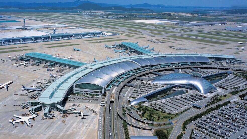 Curved airport terminal complex serving multiple parked commercial aircraft at gates, surrounded by runways, aprons, extensive parking lots, roadways and distant hills under a clear blue sky.