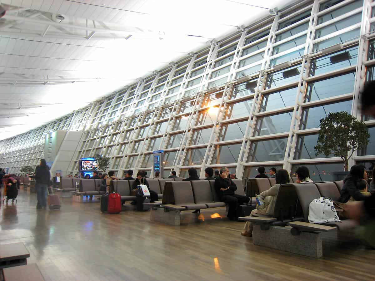 Passengers sit and wait on rows of benches, some with luggage or walking, inside a spacious airport terminal lined with tall slanted glass windows and polished wooden floors.