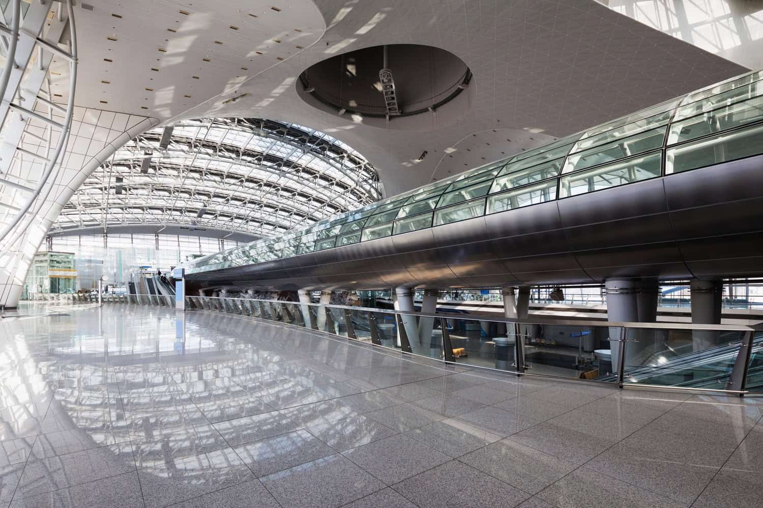 Glass-enclosed elevated walkway spans across an empty, sunlit airport concourse, supported by pillars and glass balustrades; reflective tiled floor mirrors the arched steel-and-glass ceiling and distant escalators.
