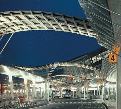 Curving metal canopy shelters waiting shuttle buses along illuminated airport terminal lanes at night; visible signage reads "4" and "5".