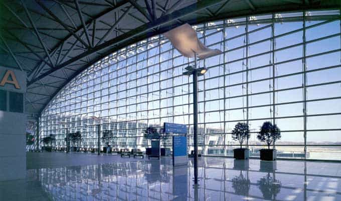 Glass-walled airport concourse curved left, sunlight streaming through grid windows and reflecting on the glossy floor; potted trees and blue signs stand by the tarmac. Text found: A