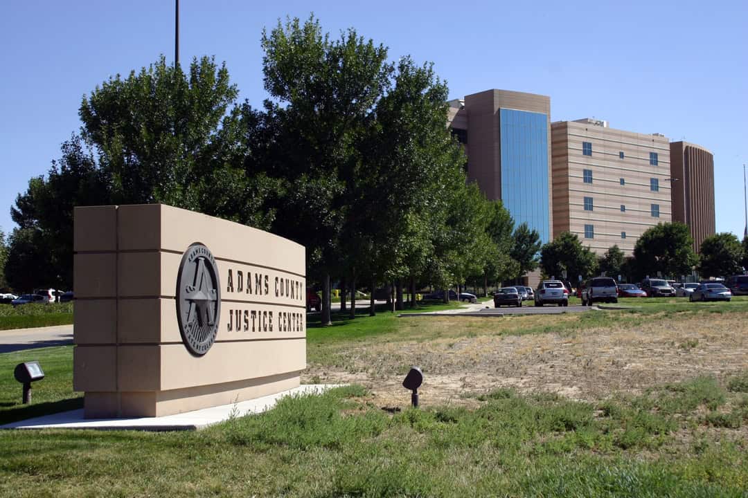 Stone sign reading "ADAMS COUNTY JUSTICE CENTER" and "ADAMS COUNTY COLORADO" stands by a grassy lawn; large multistory courthouse, trees and parked cars behind under a clear blue sky.