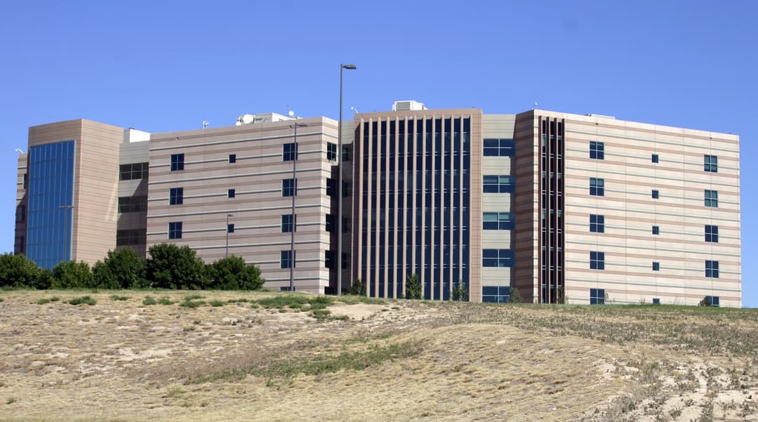 A six-story modern office building rises above a dry, sparsely vegetated slope, its beige striped facade and tall vertical glass windows reflecting sunlight under a clear blue sky.
