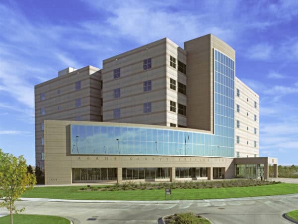 Adams County Justice Center (object) rises with large mirrored windows reflecting the blue sky (action) set behind manicured lawns and a circular driveway under clear, wispy clouds (context).

Text in image: "ADAMS COUNTY JUSTICE CENTER" and "1993"
