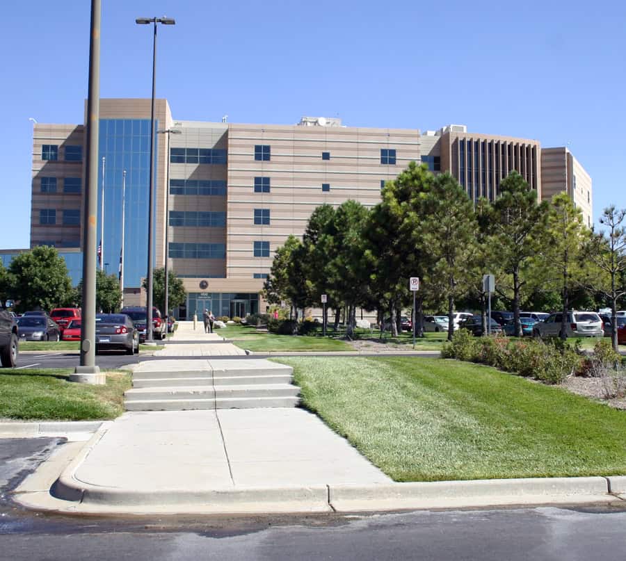 Large multi-story beige office building sits beyond a stepped concrete walkway, flanked by parked cars and pine trees, with manicured lawns and a clear blue sky overhead.