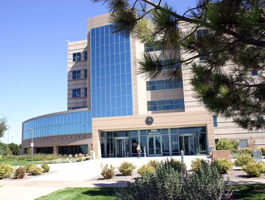 Modern tan-and-blue glass county courthouse stands fronting a paved, landscaped plaza; a person approaches the glass main entrance beneath a pine tree and clear blue sky. Text: "1100 Judicial Center Drive"; "COUNTY JUSTICE"