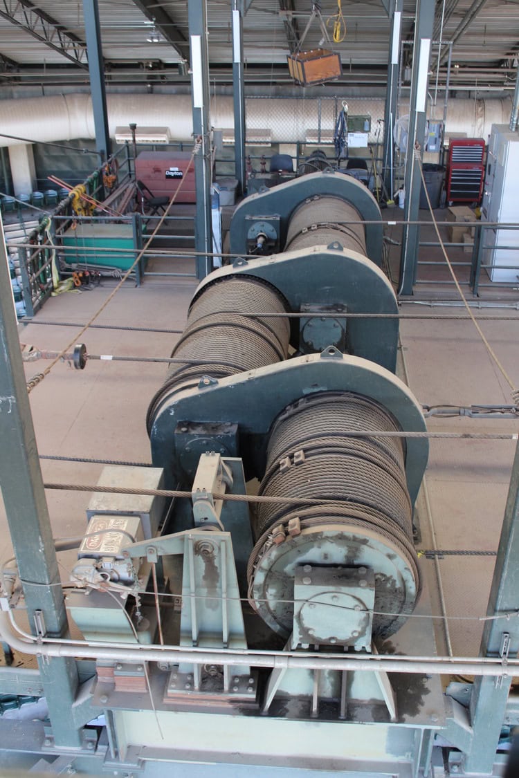 Three large industrial winch drums spooling heavy steel cable on a raised factory platform surrounded by railings, tool cabinets, and overhead hoist equipment. Text visible: "Dayton".