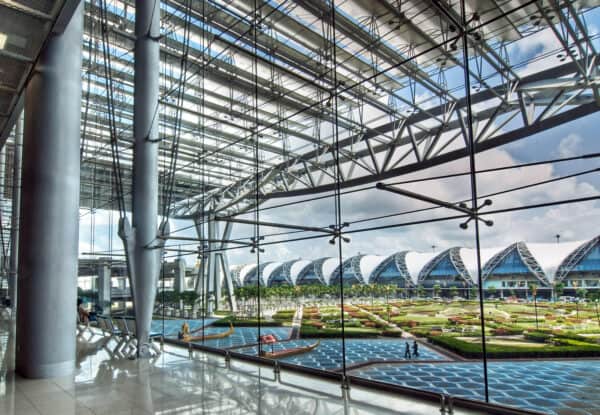 Floor-to-ceiling glass wall frames arched terminal roofs and patterned reflecting pools, supported by exposed steel trusses and columns inside a bright, spacious airport concourse with seating.