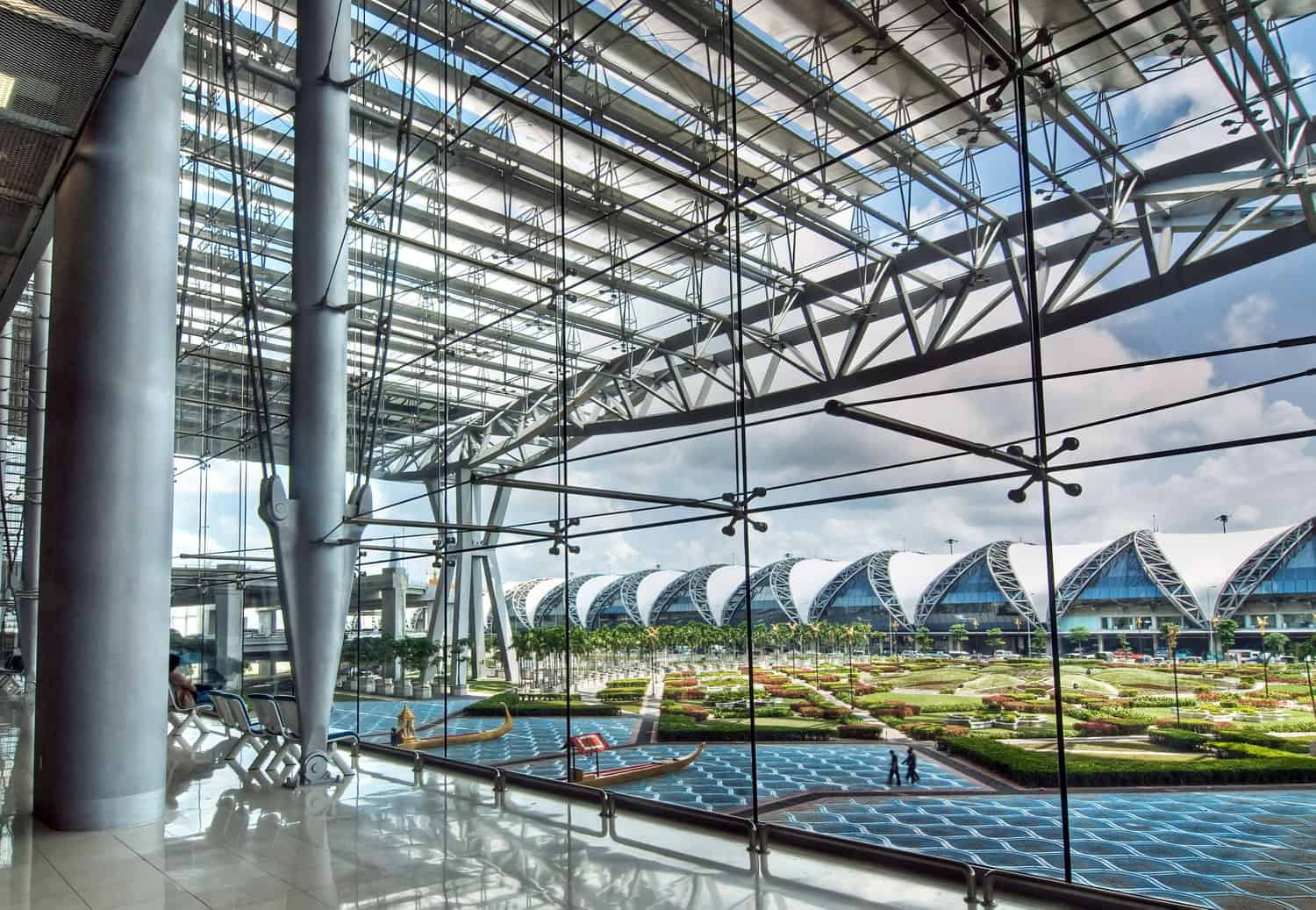 Floor-to-ceiling glass wall frames arched terminal roofs and patterned reflecting pools, supported by exposed steel trusses and columns inside a bright, spacious airport concourse with seating.