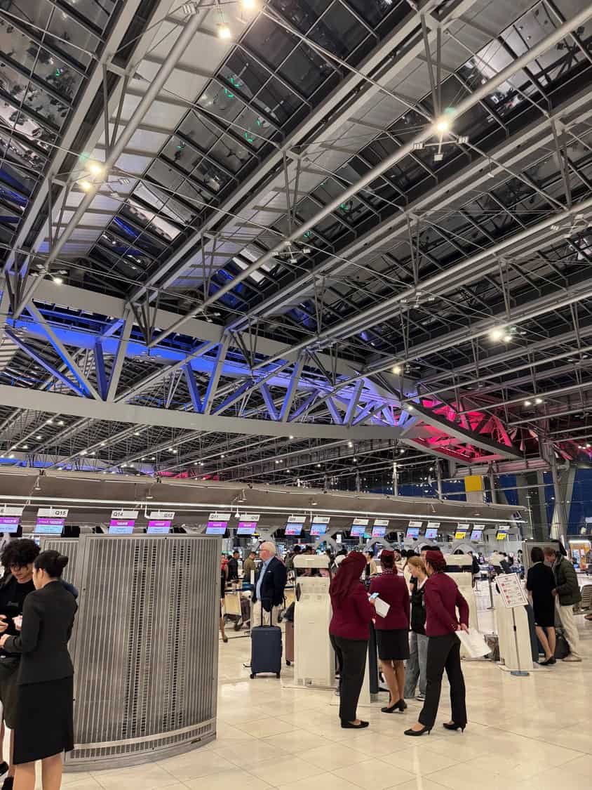 Check-in counters: airline staff assist passengers under a vast glass-and-steel ceiling lit blue and red, with rows of overhead monitors and queue kiosks in a busy terminal.

Visible text on overhead screens: Q15 Q14 Q13 Q12 Q11 Q10 Q09 Q08