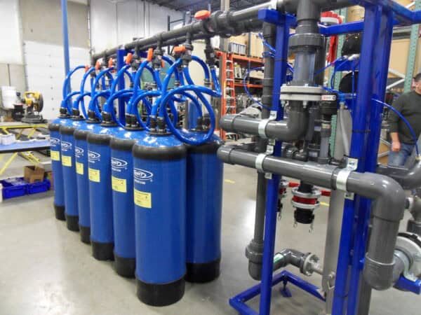 Blue cylindrical water-filter tanks lined up, connected by curved blue hoses to an overhead manifold, inside a warehouse workshop with PVC piping, shelving, workbenches, and a partially visible worker.

Text in image: "WRT" (logo) and "WRTPRODUCTS.COM" on tanks; small yellow safety labels contain additional printed warnings too small to read.