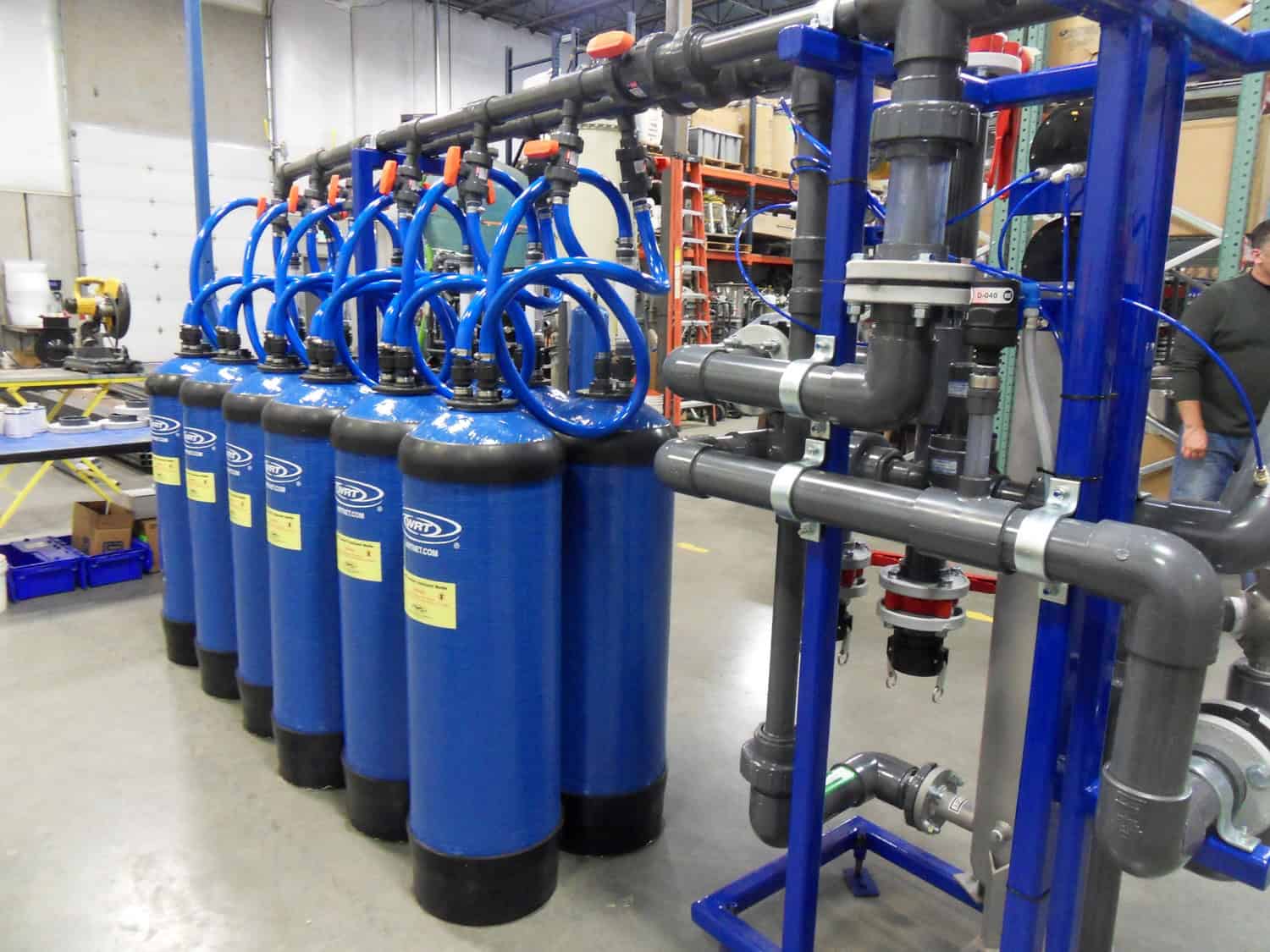 Blue cylindrical water-filter tanks lined up, connected by curved blue hoses to an overhead manifold, inside a warehouse workshop with PVC piping, shelving, workbenches, and a partially visible worker.

Text in image: "WRT" (logo) and "WRTPRODUCTS.COM" on tanks; small yellow safety labels contain additional printed warnings too small to read.