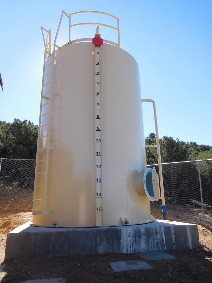 Cylindrical beige storage tank shows vertical level gauge marked "2 3 4 5 6 7 8 9 10 11 12 13 14 15", fitted with ladder and side outlet; mounted on concrete pad inside chain-link fence under clear blue sky.