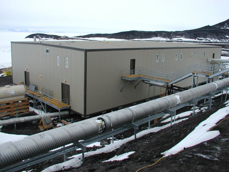 Beige modular industrial building perched on steel supports, with external staircases and platforms, adjacent to a large insulated pipeline running over snowy, rocky ground toward a distant coastal, hilly shoreline.