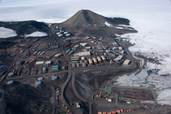 Research station complex rests at the base of a conical mountain, bordered by fragmented sea ice; roads, fuel tanks and rows of colorful buildings spread across rocky, snow-patched coastal terrain.