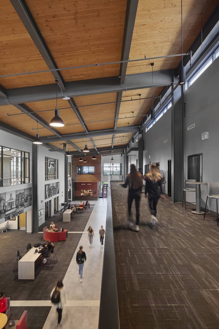 Raised carpeted walkway (object) with two blurred students walking past (action); people stroll below along a central corridor in a modern campus atrium with wooden ceiling, pendant lights, seating areas (context). Text: "THE OUTPOST".