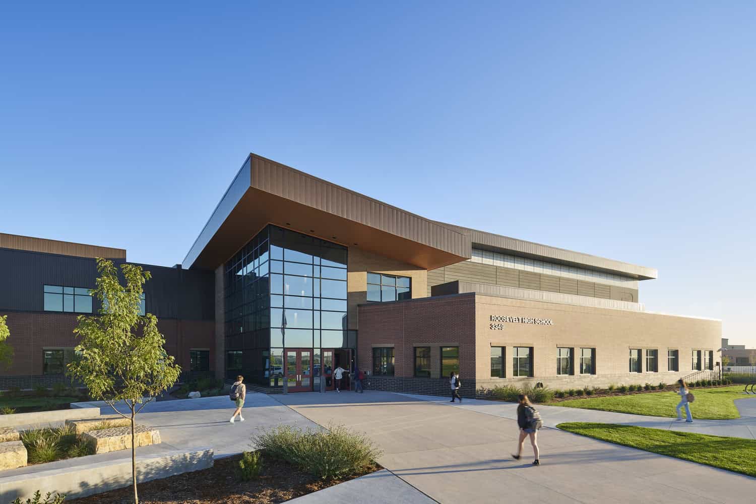 Roosevelt High School building (sign: ROOSEVELT HIGH SCHOOL 3349), sitting in late sunlight as students walk toward its tall glass entrance, on a landscaped concrete plaza with lawns and young trees.
