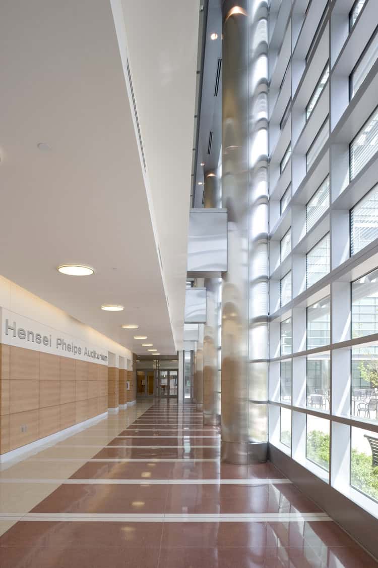 Polished lobby corridor stretches toward distant glass doors; tall stainless-steel columns and floor-to-ceiling windows reflect daylight beside a light-wood wall. Text on wall: "Hensel Phelps Auditorium"
