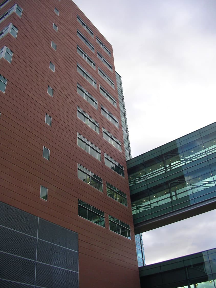 Red-brown office tower rises, its rows of windows punctuating the facade; a glass-enclosed skybridge connects it to a neighboring building under an overcast gray sky.