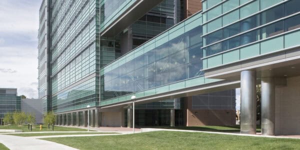 A glass-clad office building spans a suspended glass skybridge connecting sections, overlooking lawns, concrete walkways, lamp posts and stainless-steel columns under a partly cloudy sky.