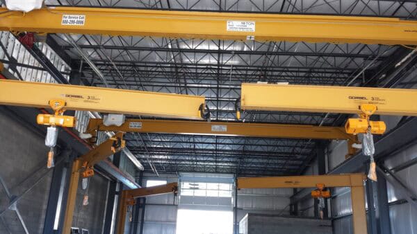 Yellow overhead cranes suspend hoist hooks across an industrial warehouse with a steel-truss ceiling and concrete-block walls. 

Text visible: "For Service Call 888-299-0896"; "GORBEL 3 TON CAPACITY"; "10 TON"; "15 TON".