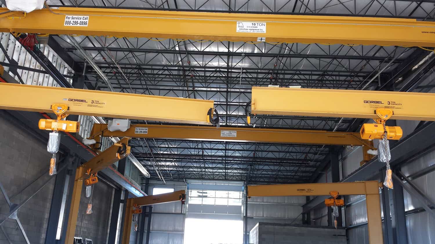 Yellow overhead cranes suspend hoist hooks across an industrial warehouse with a steel-truss ceiling and concrete-block walls. 

Text visible: "For Service Call 888-299-0896"; "GORBEL 3 TON CAPACITY"; "10 TON"; "15 TON".