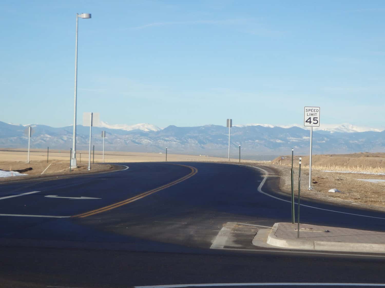 Two-lane asphalt road curves right toward distant snow-capped mountains under a clear blue sky, bordered by flat fields, lamp posts, and a speed-limit sign reading "SPEED LIMIT 45".