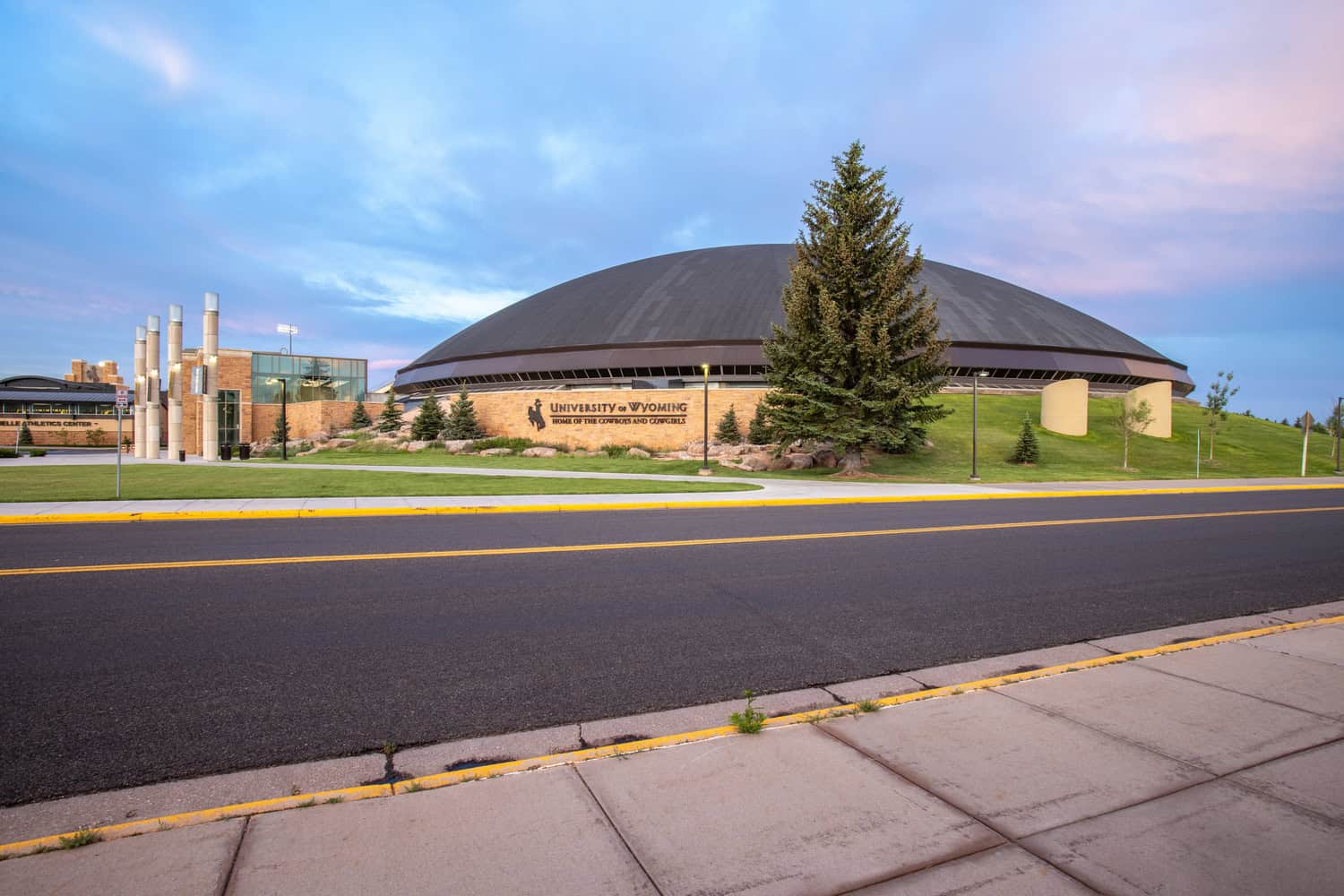 Domed arena (University of Wyoming) sits beside a large evergreen, facing a paved road and grassy campus at dusk.

Visible text (transcribed):
- "UNIVERSITY of WYOMING"
- "HOME OF THE COWBOYS AND COWGIRLS"
- partially visible: "ATHLETICS CENTER"