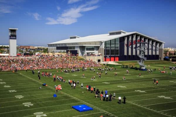 Football team practicing — players run drills on a marked practice field — packed crowd on a grassy hill beside a large training facility with Broncos logo beneath a clear blue sky. Text: "UCHealth"