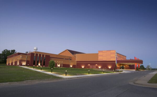 Large red-orange civic building glows with exterior lights, its curved canopy entrance welcoming vehicles; illuminated walkway lights edge a grassy lawn beside a road beneath a clear twilight sky.