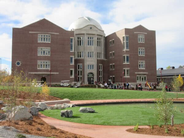 Large red-brick academic building with central stone dome stands behind a green lawn; people gather near the entrance while a curved brick path and reflecting pond with rocks fill the foreground.