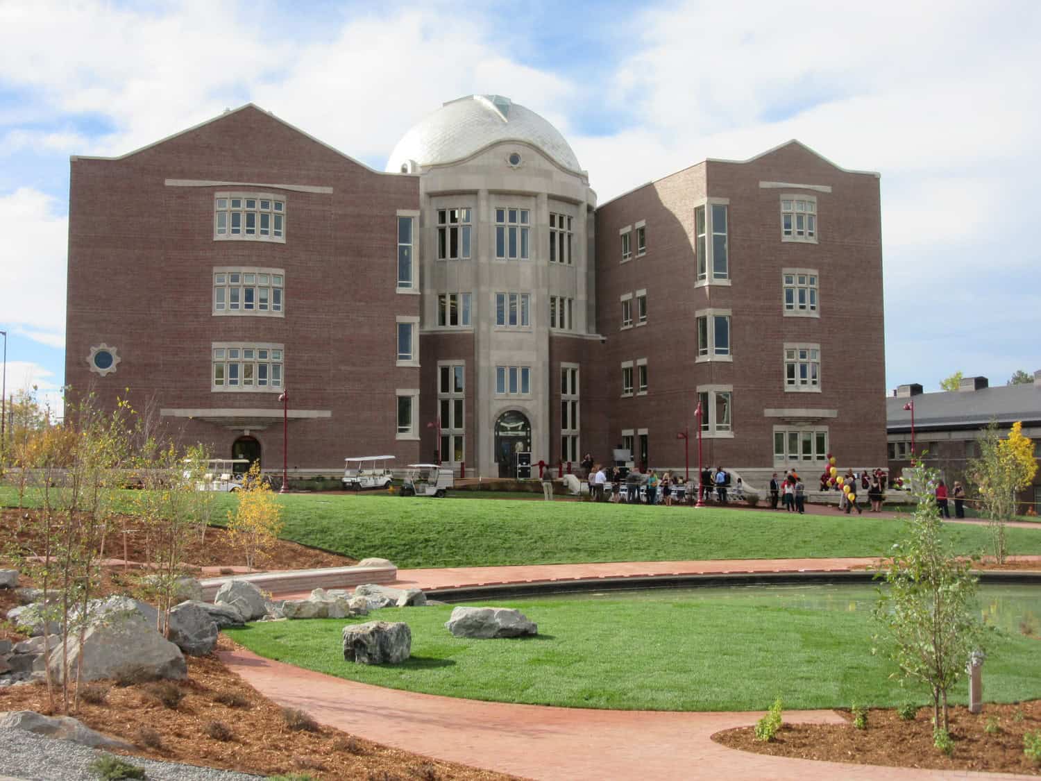 Large red-brick academic building with central stone dome stands behind a green lawn; people gather near the entrance while a curved brick path and reflecting pond with rocks fill the foreground.