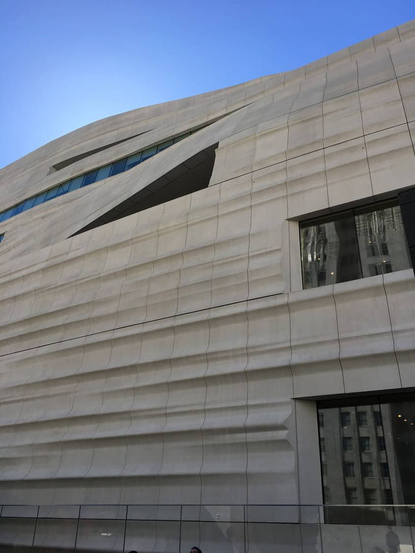 Sculpted concrete building facade undulates with horizontal ribs and angular slit windows, rising diagonally against a clear blue sky, reflective glass showing neighboring buildings at street level.