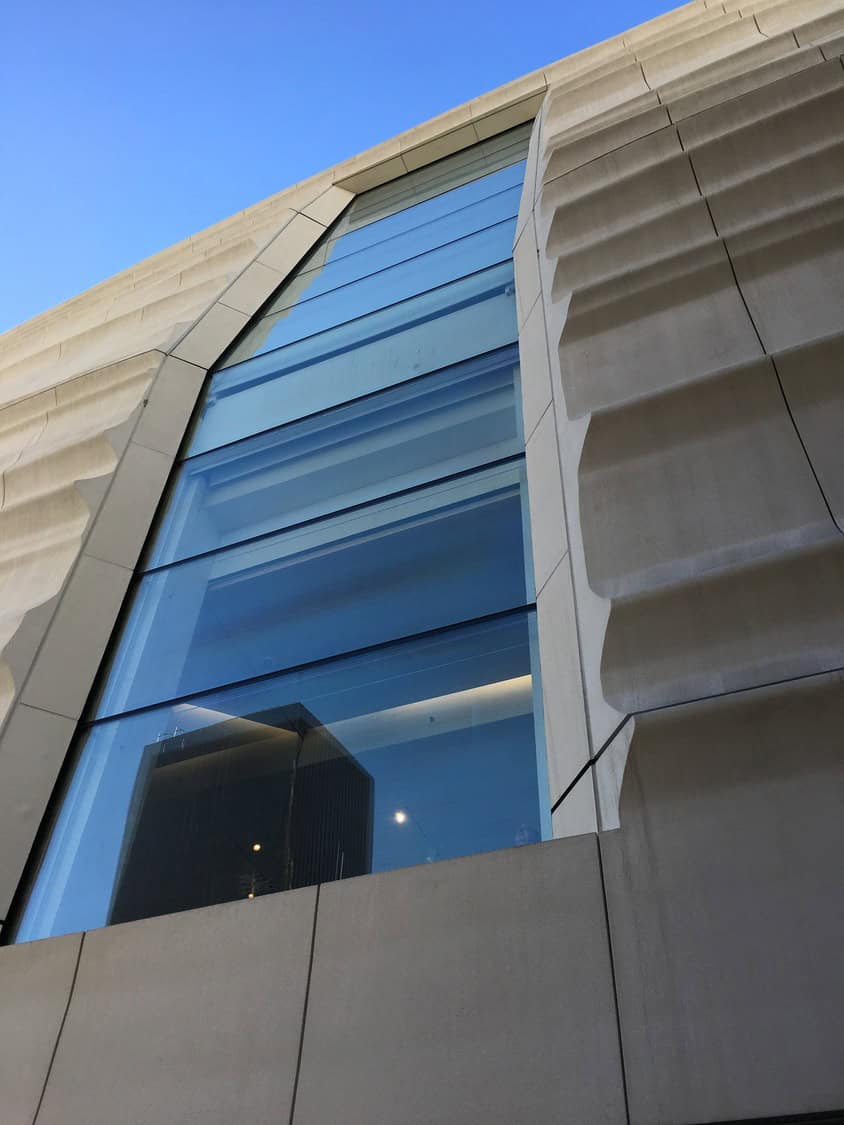 A tall blue-tinted glass window reflects a nearby dark skyscraper and interior lights, set within sculpted concrete panels with vertical grooves, viewed from ground level against a clear blue sky.