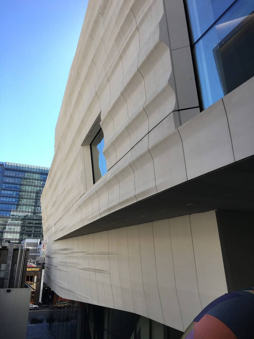Wavy limestone building facade curves outward, casting rhythmic shadows along an urban block with glass office tower and clear blue sky; small street sign reads "BREWERY".