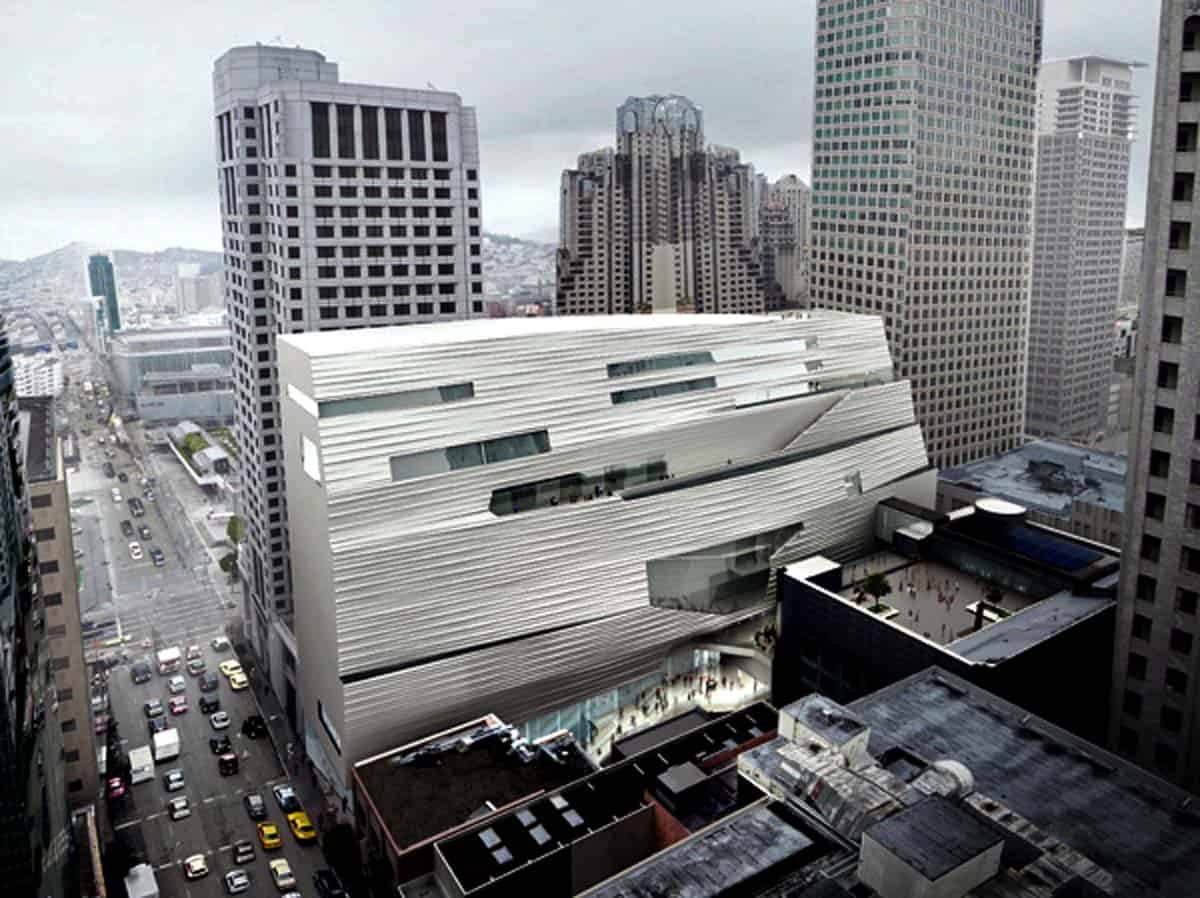 Large angular white building occupies a downtown block, its ribbed slanted facade pierced by long horizontal windows and terraces, surrounded by high-rise towers above a busy, car-filled street under overcast sky.