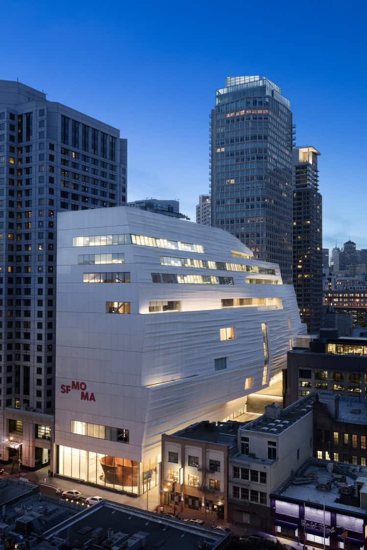 A white sculptural museum building (text: "SF MOMA") glows at dusk, occupying a city block and surrounded by tall glass office towers and narrow streets in downtown San Francisco.