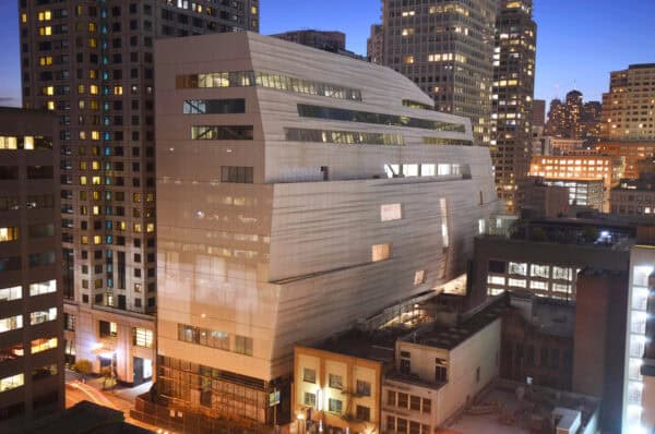 A large white curved building rises, horizontal slit windows glowing; surrounded by tall apartment and office towers at dusk, overlooking a city block with lit windows and streets.