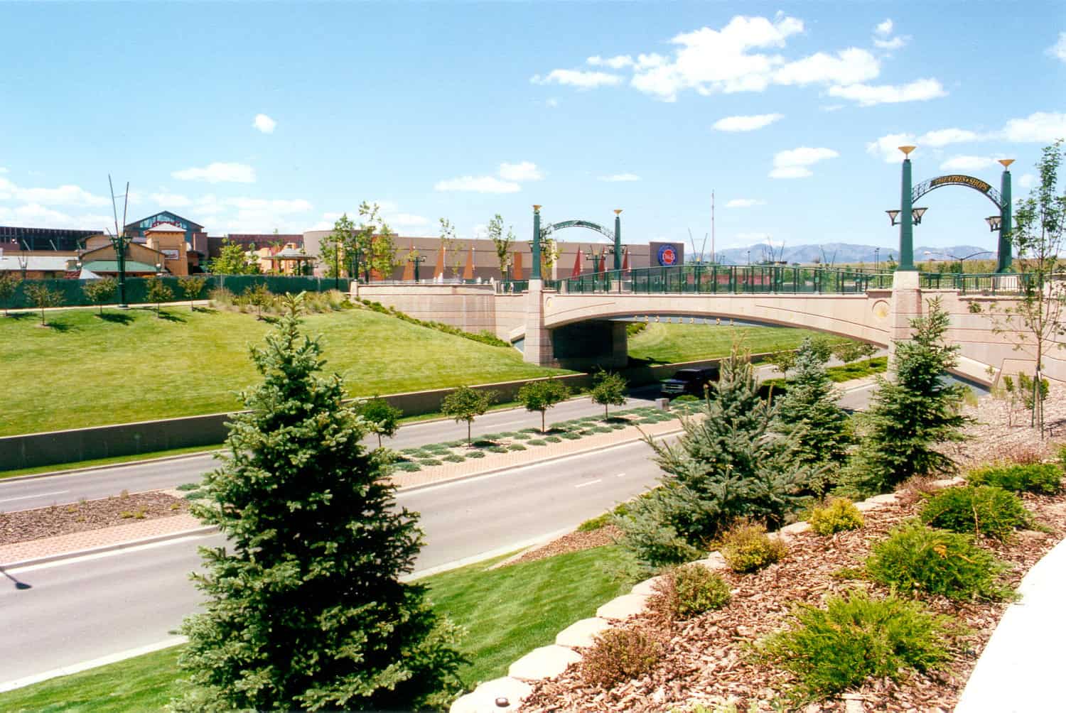 Stone pedestrian bridge spans a multi-lane road, linking a shopping complex; manicured grassy slopes, evergreen trees and landscaped beds flank the roadway beneath a bright blue sky. Text: "THEATRES & SHOPS", "D&B".