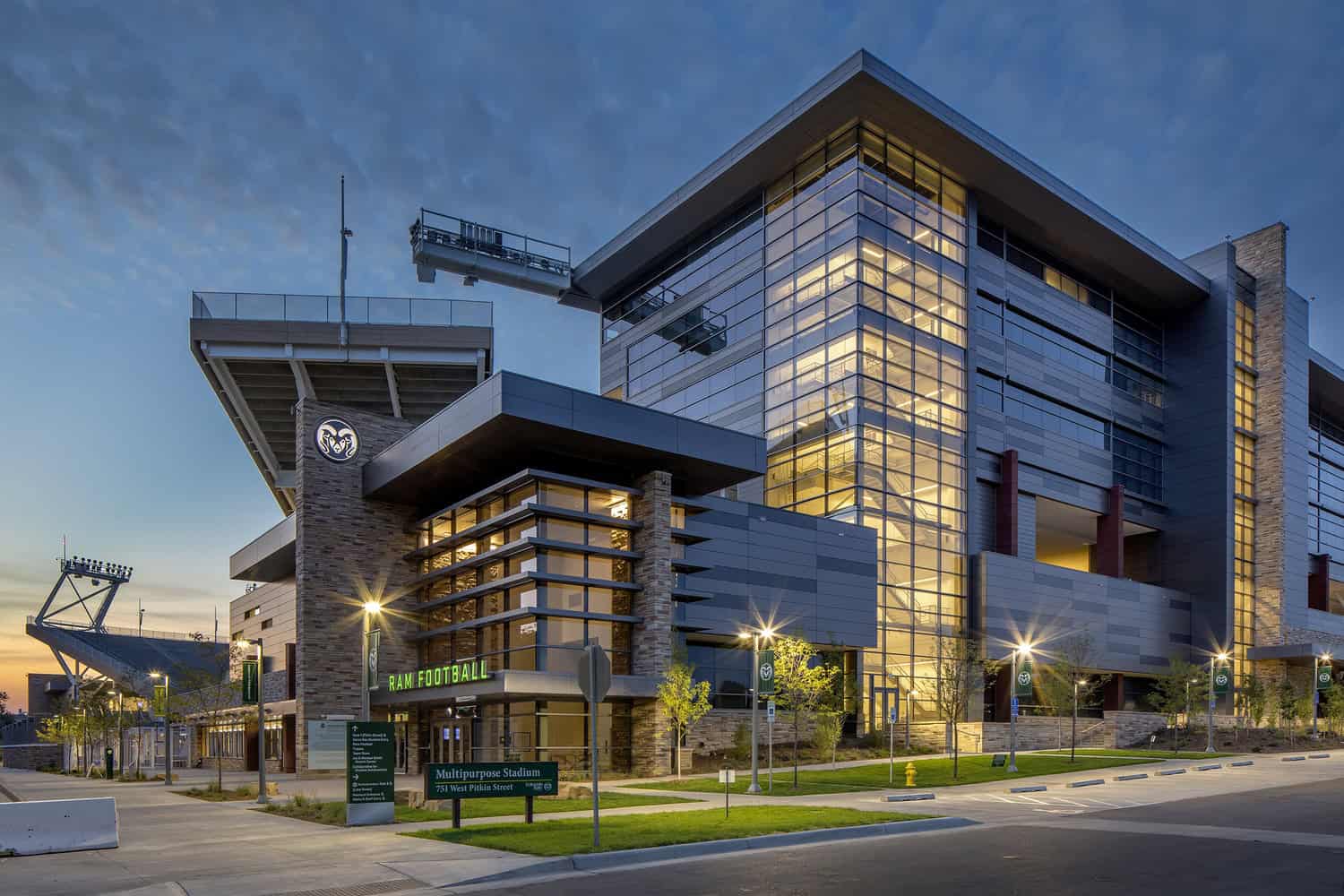 Multipurpose stadium glows at dusk, glass-and-steel façade lit from within, cantilevered seating and walkway visible; quiet street, lampposts, young trees and signage in foreground.

Text: "RAM FOOTBALL"; "Multipurpose Stadium 751 West Pitkin Street"