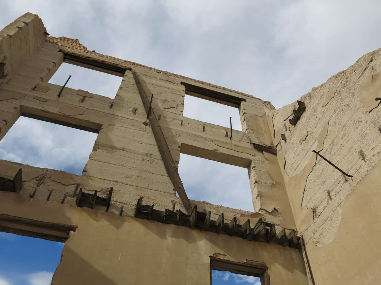 Concrete building shell reveals large rectangular window openings and exposed rebar and wooden beams, crumbling plaster and jagged edges, viewed upward against a cloudy blue sky.