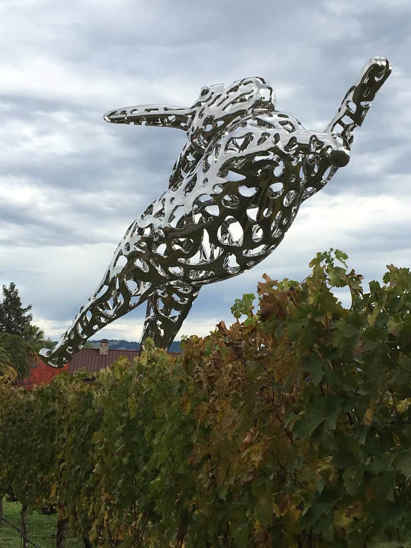 Polished, perforated stainless-steel human-like sculpture leaps forward above rows of grapevines, reflecting the cloudy sky; a vineyard with distant trees and rooftops forms the overcast rural backdrop.