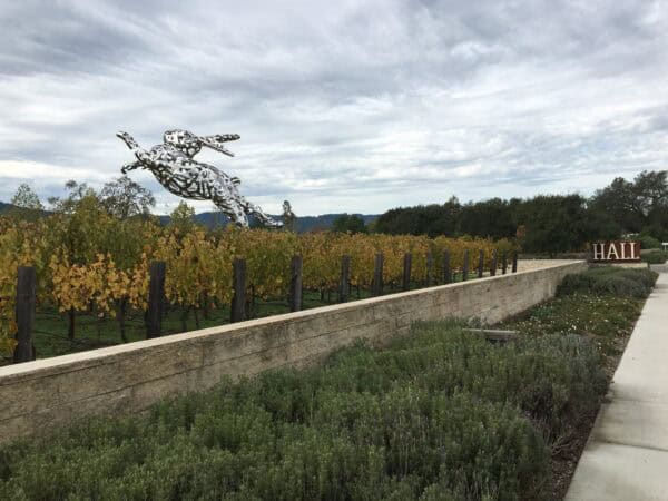 A large perforated metal rabbit sculpture appears to leap over rows of yellowing grapevines, set behind a low stone wall and walkway under an overcast sky; a sign reads "HALL".