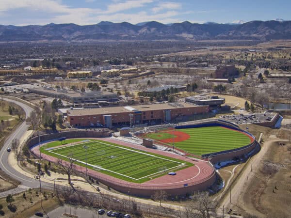A dual-sport stadium: green football field with running track sits adjacent to a baseball diamond with red infield; empty stands and surrounding school buildings, parking lots, winding roads, distant mountains.