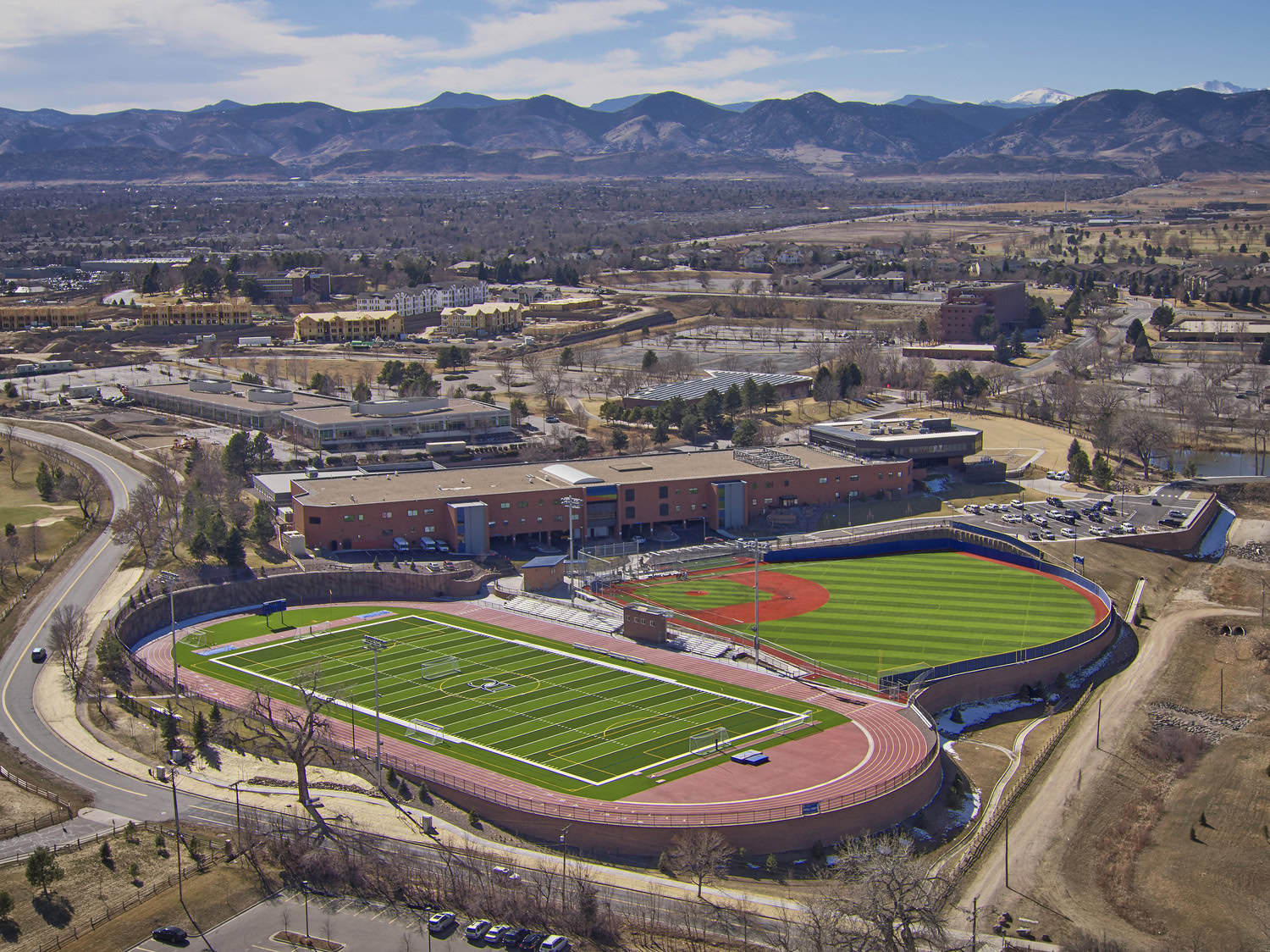 A dual-sport stadium: green football field with running track sits adjacent to a baseball diamond with red infield; empty stands and surrounding school buildings, parking lots, winding roads, distant mountains.