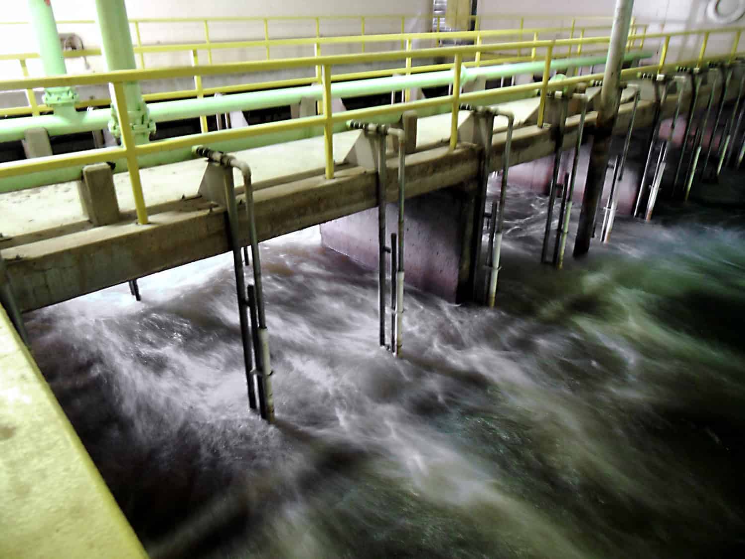 Concrete walkway with yellow railings and attached vertical metal pipes spans over churning, frothy water, inside an indoor industrial water-treatment basin with overhead green pipelines.